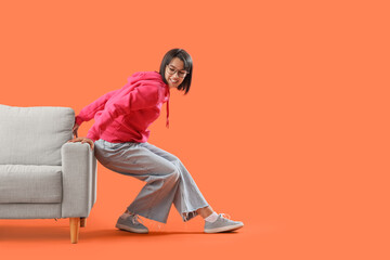 Young woman pushing sofa against orange background