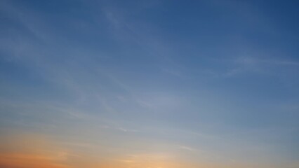 A tranquil sunset with a clear blue sky gently transitioning to warm hues of orange and yellow near the horizon. Delicate wispy clouds are scattered across the sky. Sunset sky background.
