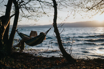 A lovely young girl enjoys a peaceful reading session in a hammock, overlooking a lake during a...