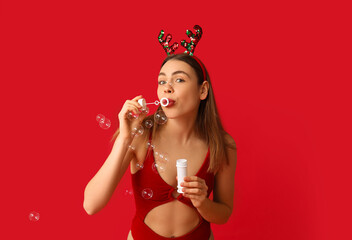 Young woman in swimsuit and reindeer horns blowing soap bubbles on red background. Christmas in July