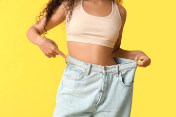 Young African-American woman pointing at loose jeans on yellow background. Weight loss concept