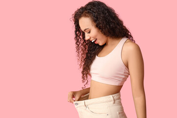 Surprised young African-American woman in loose jeans on pink background. Weight loss concept