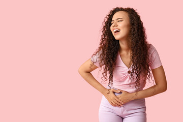 Young African-American woman with severe stomach ache screaming on pink background