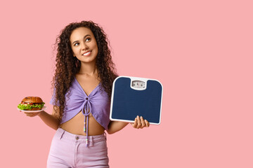 Happy young African-American woman with scales and burger on pink background. Diet concept
