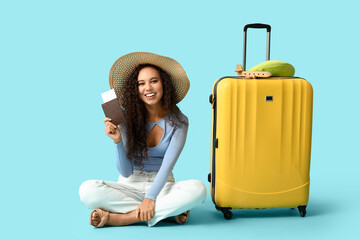 Happy young African-American woman with suitcase, passport and ticket sitting on blue background