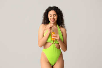 Beautiful young African-American woman in stylish green swimsuit with bottle of cocktail on grey background