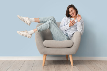 Beautiful young woman in stylish jeans with mobile phone sitting on armchair near blue wall