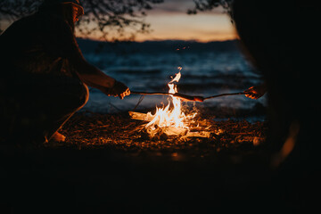 Two friends gather around a warm campfire by a lake, roasting food and enjoying each other's...