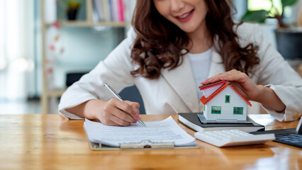 Businesswoman signing contract while holding house model, showcasing insurance or real estate...