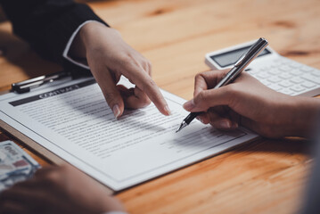 Close-up of hands signing a contract on a wooden desk, with a pen, a calculator, and documents in...