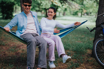 A joyful young couple relaxes in a blue hammock, laughing together in a lush urban park on a beautiful sunny day.