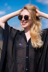 Beautiful, attractive, happy girl posing on a sandy beach on a sunny day wearing a silver swimsuit, sunglasses and a black beach tunic. Beachwear