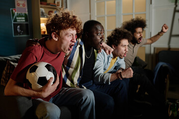 Multi-ethnic group of young male soccer fans sitting on couch in living room watching match on TV