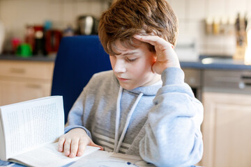 Cute blond kid boy reading book in domestic room. Excited child reading loud. Schoolkid, family,...