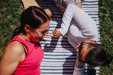 High angle view of two female friends laughing and relaxing on a striped picnic blanket in a green park on a sunny day.