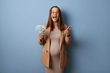 Excited amazed pregnant woman wearing beige jacket and dress holding euro banknotes and using...