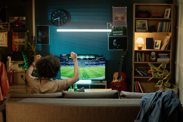 Rear view of unrecognizable biracial man sitting on sofa in living room watching soccer game match...