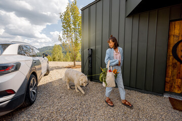 Young woman going home with her cute dog and groceries in bag arrived by electric car into her modern house. Concept of sustainable lifestyle, wellbeing and friendship with pets