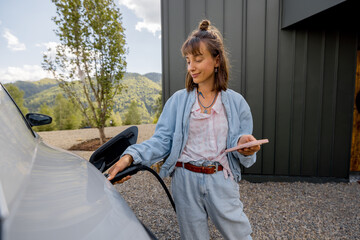 Young woman plugs a charger into electric vehicle standing with smart phone near her house...