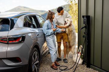 Portrait of a young couple with their cute white dog near house and modern electric car outdoors....