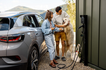 Portrait of a young couple with their cute white dog near house and modern electric car outdoors. Concept of family and wellbeing