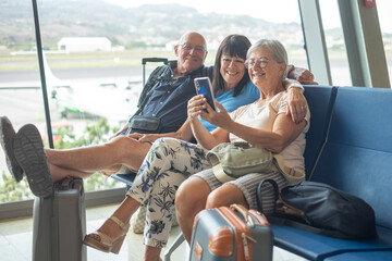 Smiling group of senior friends sharing tech and social on mobile phone sitting in airport departure area waiting for boarding. Travel and tourism concept, retiree lifestyle