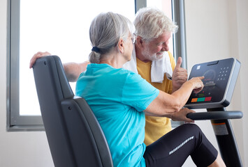Athletic senior couple enjoying stay fit working together in gym, woman doing exercises on stationary bike while talks with elderly man showing goals