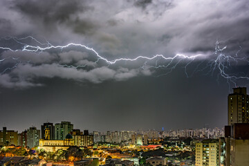 Color photo of a lightning storm over a cityscape at night. Bolts illuminate dark clouds and buildings, creating an intense atmosphere, showcasing nature's power.