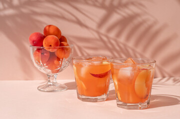 Healthy summer fruit nutrition, apricots and peaches in vase and glasses with fresh peach fruit juice with ice on bright peachy pink background with sunlight floral shadows