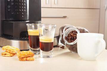 Two glass shorts of espresso near coffee maker, cookies and cans of coffee beans on light table.