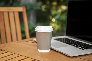 A paper cup of coffee and a laptop on a wooden table on a blurred forest background, work in...