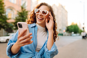 Picture of pretty young woman staying on the street holding phone in hands. Blogging, technology,...