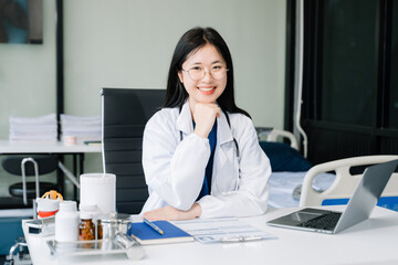 Confident young female doctor in white medical uniform sit at desk working on computer. Smiling use laptop write in medical journal