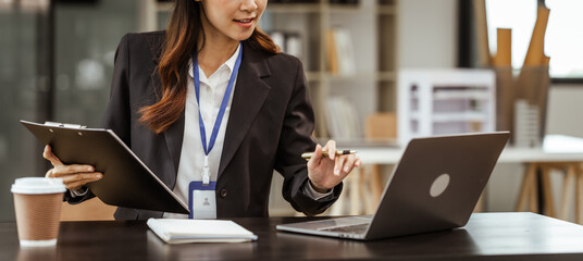 A young Asian businesswoman is seen diligently working at her desk. She's immersed in tasks ranging...