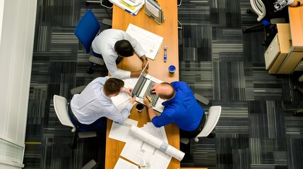 Three business professionals collaborating on blueprints and using a laptop in a modern office setting, viewed from above.