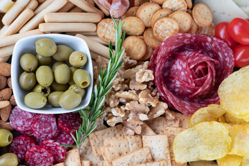 Assortment of savory snacks perfect for sharing, display of crackers, breadsticks, potatoes chips around a salami slices arrangement complimented by cherry tomatoes, walnuts and a bowl of green olives