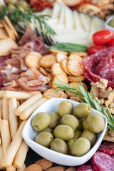 Charcuterie board featuring a ceramic bowl filled with green olives set in the middle of large selection of delicacies: cured meats, slices of cheese, breadsticks, crackers, cherry tomatoes and nuts