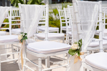 Close-up of an elegant seating arrangement for a wedding or a romantic ceremony featuring white chairs and cushions, draped in sheer fabric and set in place with rose bouquets tied with golden ribbons