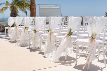 Elegant setup of chairs wrapped in sheer fabric tied with delicate golden ribbons and fresh bouquets of white roses and greenery, concept for dream destination, perfect location for romantic elopement