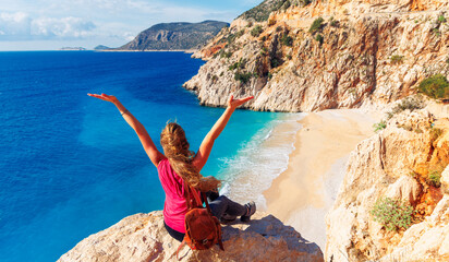 Happy young female sitting on cliff rock with arms raised enjoying beautiful beach- Turkey, Antalya