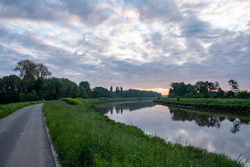A path stretching towards a river, framed by lush greenery and trees under a cloudy sky, showcasing a serene natural landscape