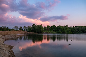 The image shows a serene lake reflecting the colors of a sunset, with trees silhouetted in the foreground under a sky filled with dramatic clouds