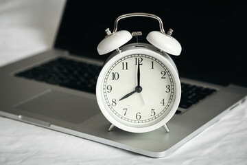 White alarm clock and laptop in a bed, work from home on white sheets in bedroom.