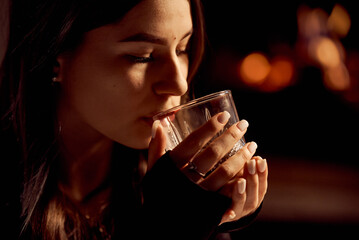A beautiful young girl drinks from a glass of alcohol in the evening.