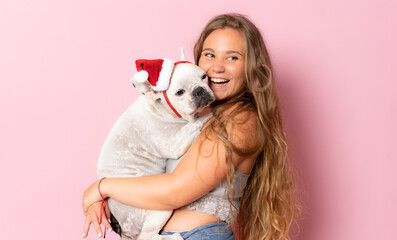 Happy young woman playing with french bulldog in santa hat on pink background.