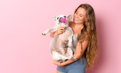 Cute young woman kisses and hugs her french bulldog dog isolated on pink background.