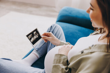 Cropped young pregnant woman with belly wear casual clothes hold ultrasound image sit on blue sofa...