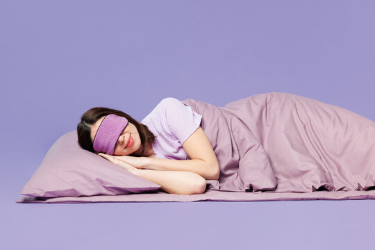 Side profile view young calm woman wears pyjamas jam cover eyes with sleep eye mask rest relax at home lay down on bed isolated on plain pastel purple background studio. Good mood night nap concept.