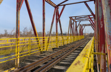 Yellow railings, high beams, old rusty rails, brown wooden sleepers of a railway bridge. Blue sky