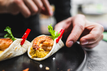 Close-up of a hand preparing a shrimp taco with fresh ingredients on a black plate, showcasing culinary skills and fresh produce.